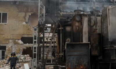 Worker in protective jacket stands beside rusty industrial machinery in a ruined factory building.