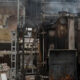 Worker in protective jacket stands beside rusty industrial machinery in a ruined factory building.