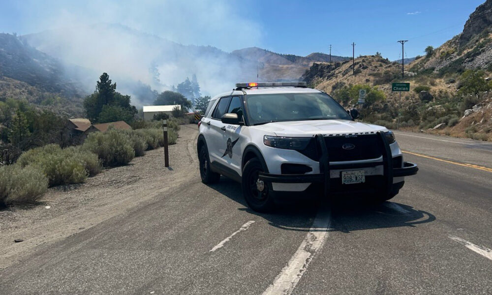 White police SUV with lights on blocking a mountain highway as smoke from wildfires rises in the background, suggesting road closure and firefighting activity.