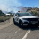 White police SUV with lights on blocking a mountain highway as smoke from wildfires rises in the background, suggesting road closure and firefighting activity.