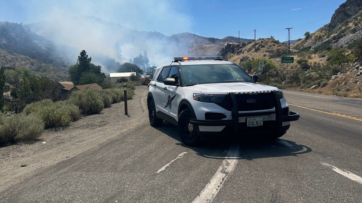 White police SUV with lights on blocking a mountain highway as smoke from wildfires rises in the background, suggesting road closure and firefighting activity.