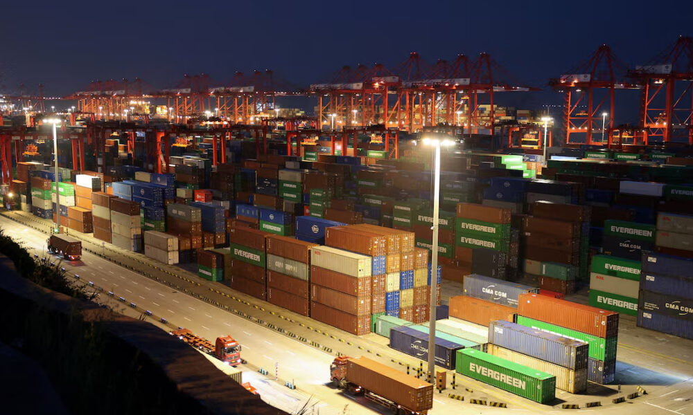 Night view of a shipping port with red gantry cranes and stacked colorful cargo containers under bright yard lights at dusk.