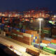 Night view of a shipping port with red gantry cranes and stacked colorful cargo containers under bright yard lights at dusk.