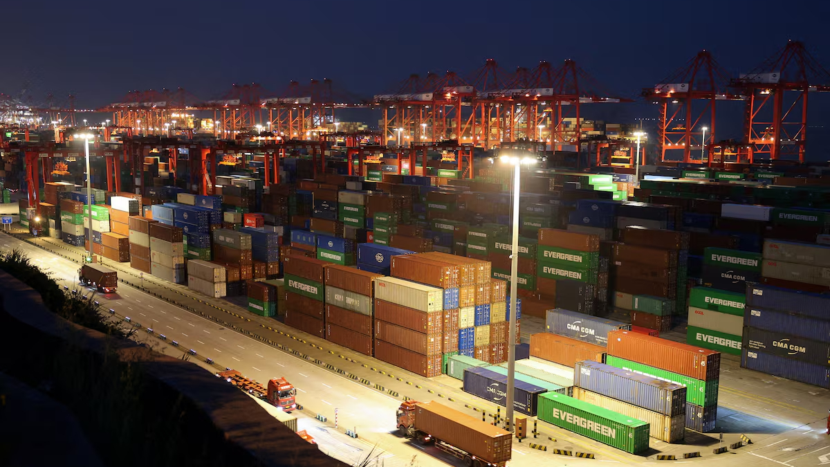 Night view of a shipping port with red gantry cranes and stacked colorful cargo containers under bright yard lights at dusk.