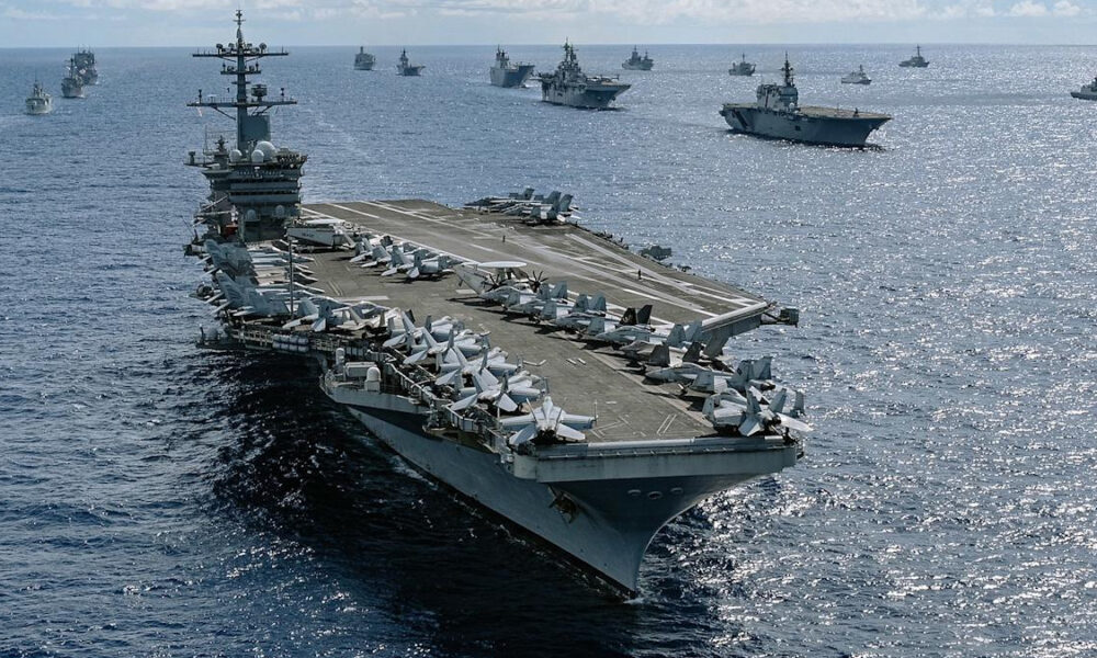 A large aircraft carrier sails on the open sea with fighters lined up on its flight deck and multiple ships in the distance.