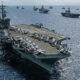 A large aircraft carrier sails on the open sea with fighters lined up on its flight deck and multiple ships in the distance.