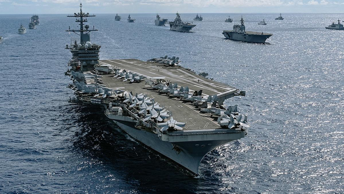 A large aircraft carrier sails on the open sea with fighters lined up on its flight deck and multiple ships in the distance.