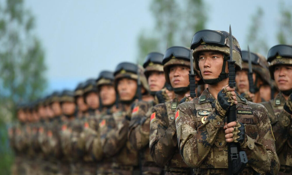 Soldiers in camouflage uniforms and helmets standing in formation, holding rifles at attention.