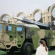 Camouflage military missile launcher truck on display with a mounted rocket and a spectator in foreground.