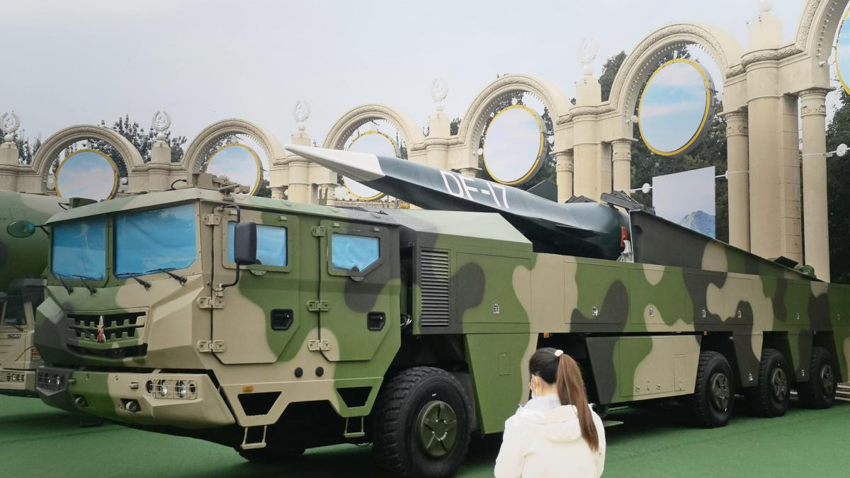 Camouflage military missile launcher truck on display with a mounted rocket and a spectator in foreground.