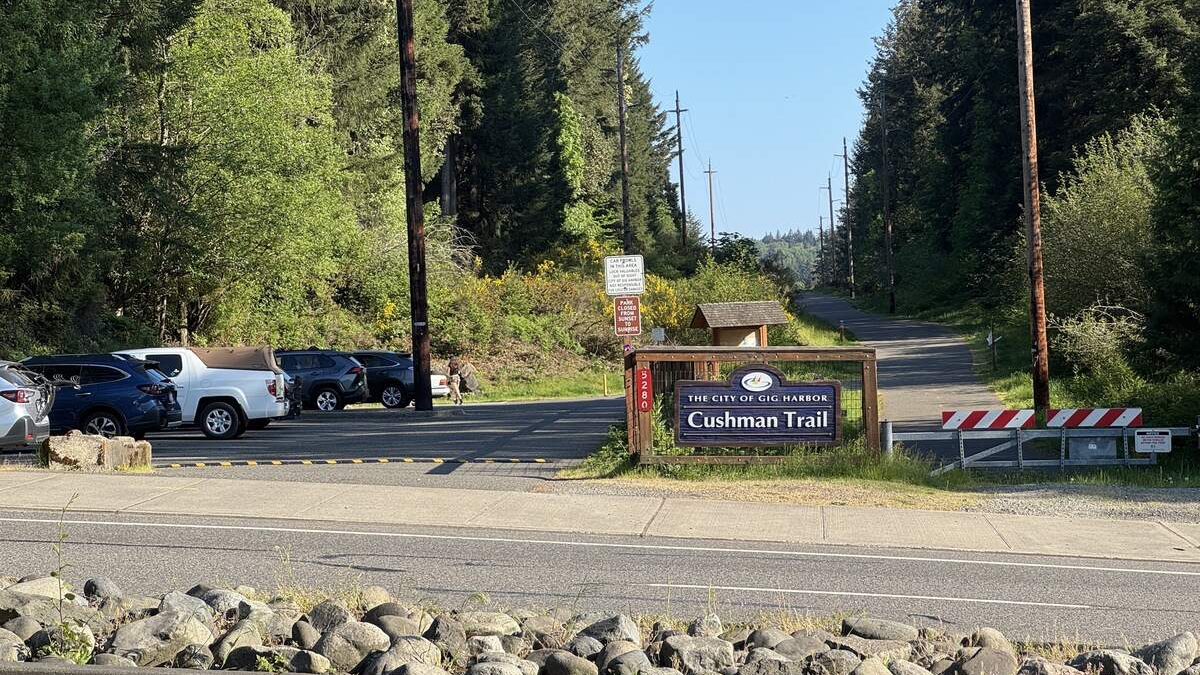 Entrance to Cushman Trail in Gig Harbor with a wooden sign, a row of parked cars, and a tree-lined road ahead.