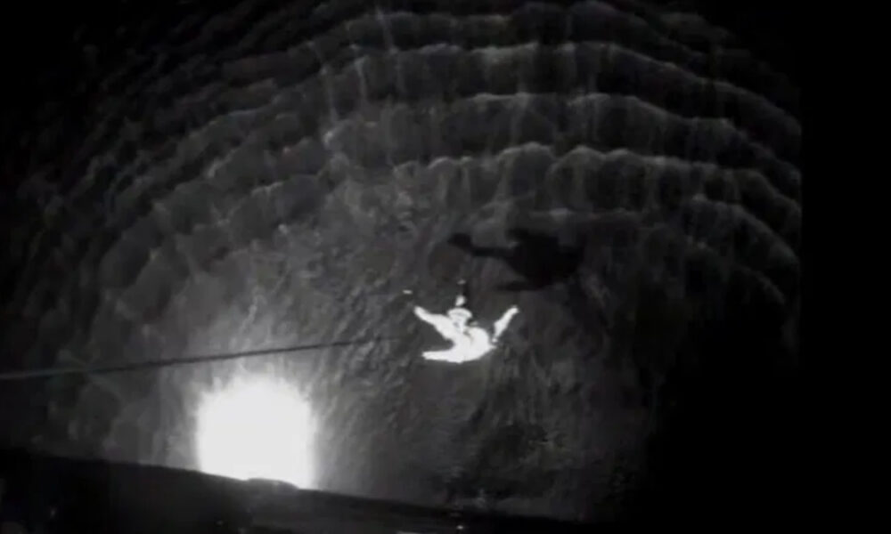 Person suspended mid-air inside a dark circular shaft, held up by a rope or cable against the stone walls at night