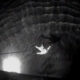 Person suspended mid-air inside a dark circular shaft, held up by a rope or cable against the stone walls at night