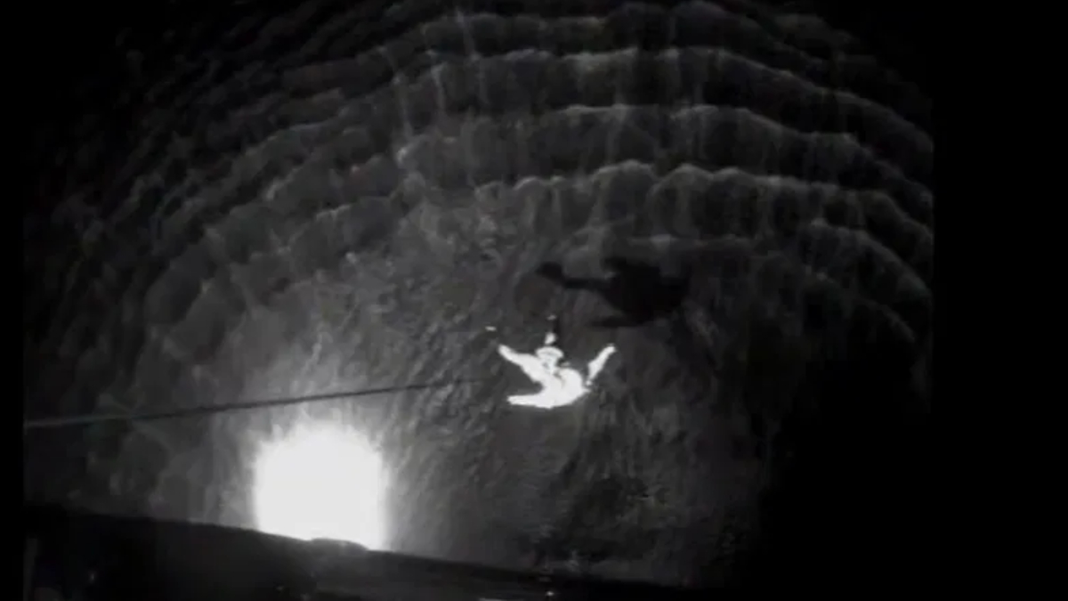 Person suspended mid-air inside a dark circular shaft, held up by a rope or cable against the stone walls at night