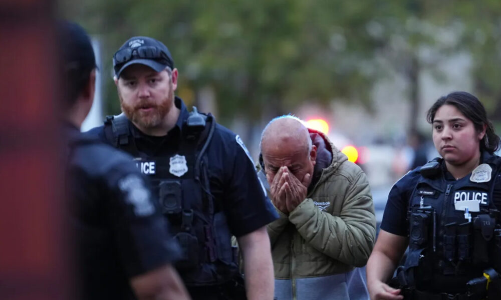 Distressed man in a hoodie covers his face with his hands as two police officers stand on either side outdoors at dusk, blurred red/blue emergency lights in background.