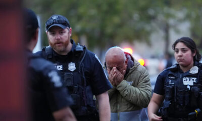 Distressed man in a hoodie covers his face with his hands as two police officers stand on either side outdoors at dusk, blurred red/blue emergency lights in background.