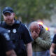 Distressed man in a hoodie covers his face with his hands as two police officers stand on either side outdoors at dusk, blurred red/blue emergency lights in background.