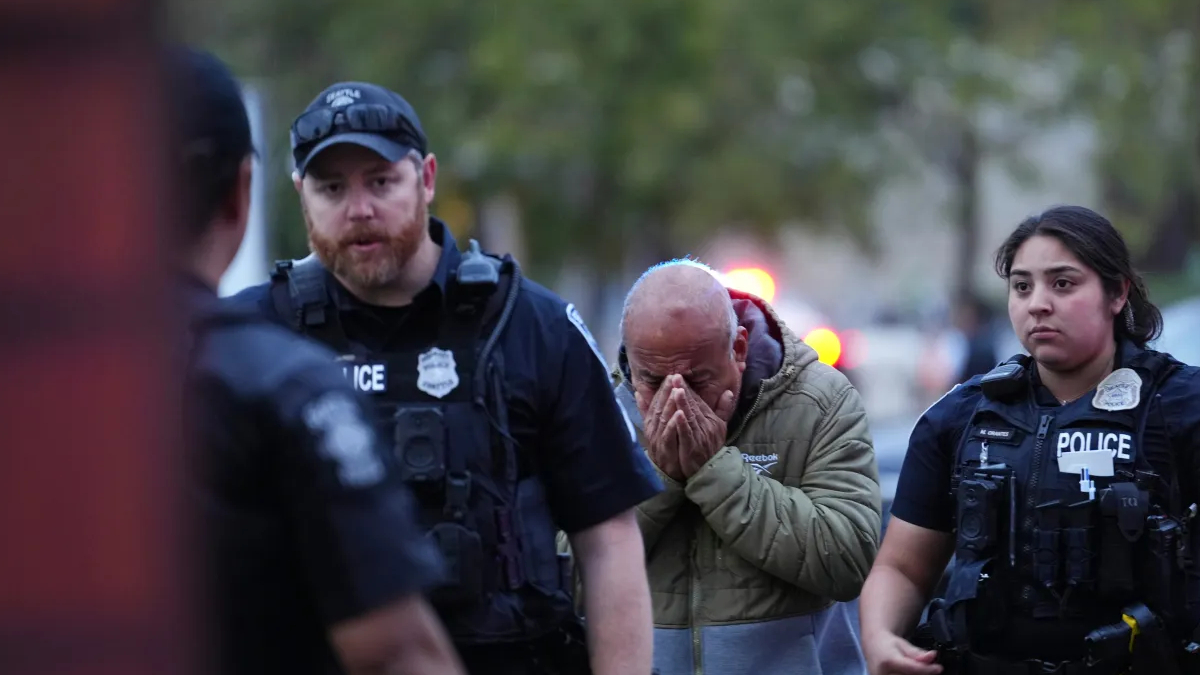 Distressed man in a hoodie covers his face with his hands as two police officers stand on either side outdoors at dusk, blurred red/blue emergency lights in background.