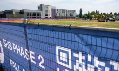 Blue construction fence banner reading 'SHS Phase 2' with a modern building and active construction equipment in the background.