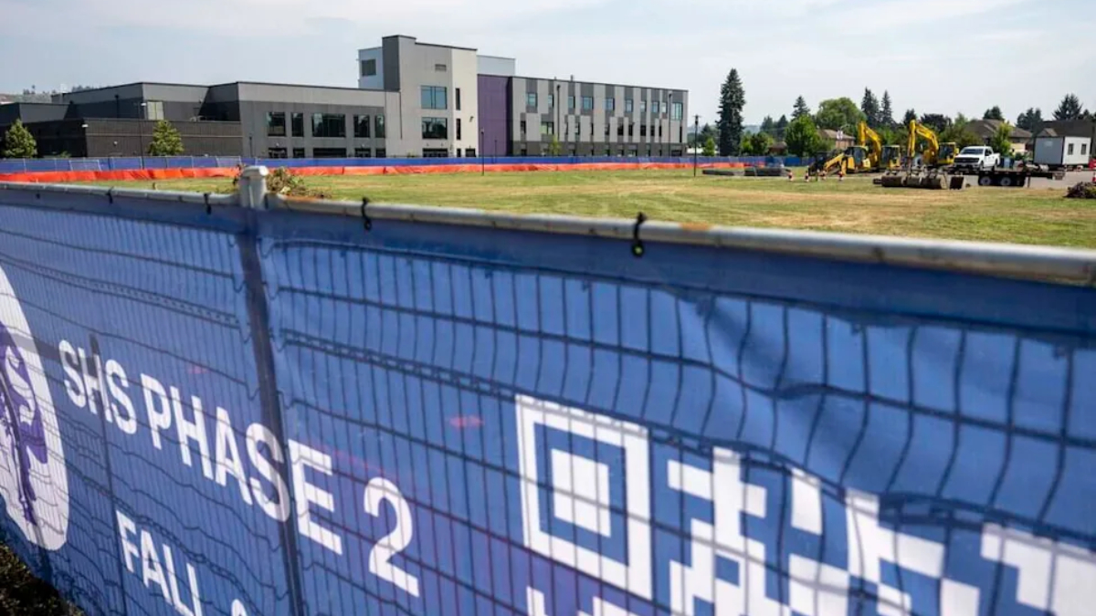 Blue construction fence banner reading 'SHS Phase 2' with a modern building and active construction equipment in the background.