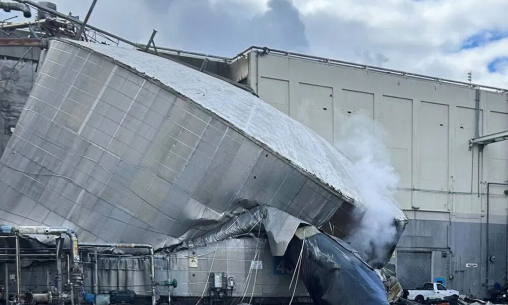 Collapsed large metallic ductwork leaning against a building after an industrial accident, with steam or smoke nearby.