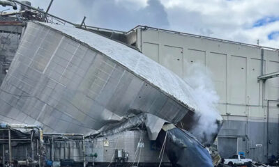 Collapsed large metallic ductwork leaning against a building after an industrial accident, with steam or smoke nearby.