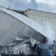 Collapsed large metallic ductwork leaning against a building after an industrial accident, with steam or smoke nearby.