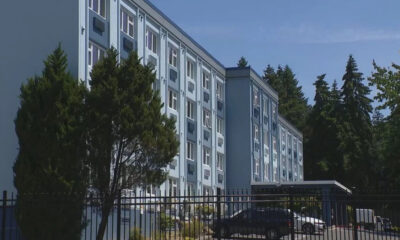 Blue multi-story apartment building with rows of windows, a gated parking area, and tall trees nearby in a sunny setting with a clear sky.