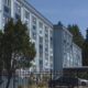 Blue multi-story apartment building with rows of windows, a gated parking area, and tall trees nearby in a sunny setting with a clear sky.