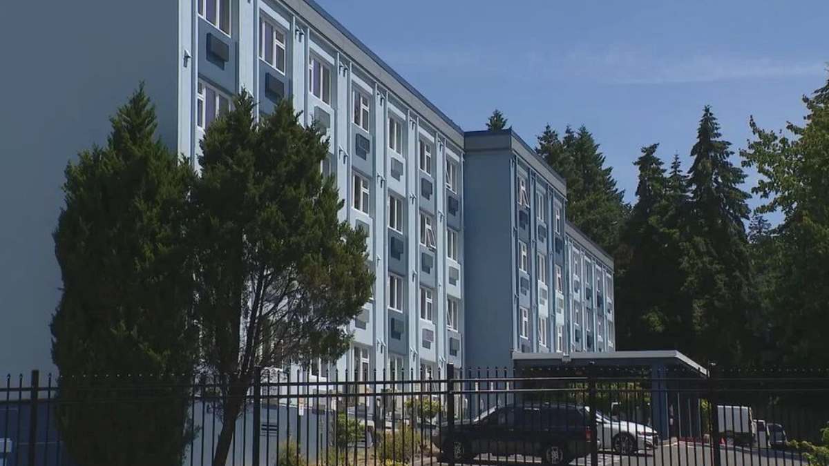 Blue multi-story apartment building with rows of windows, a gated parking area, and tall trees nearby in a sunny setting with a clear sky.