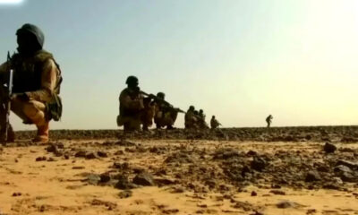 Soldiers in desert camouflage kneeling with rifles, lined up along a rocky, sunlit landscape.