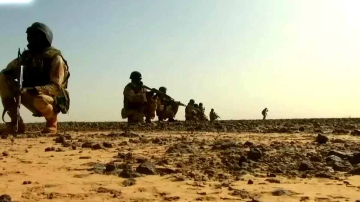 Soldiers in desert camouflage kneeling with rifles, lined up along a rocky, sunlit landscape.
