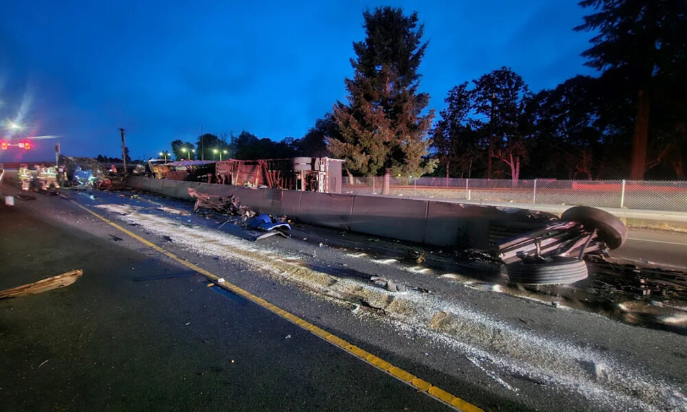 Wrecked truck and debris across a highway at dusk, emergency responders in the distance, damaged guardrail and overturned trailer on the right side.