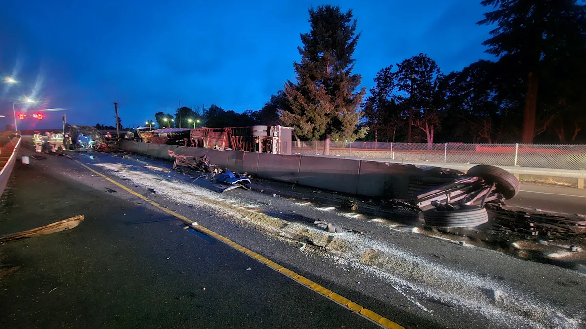Wrecked truck and debris across a highway at dusk, emergency responders in the distance, damaged guardrail and overturned trailer on the right side.
