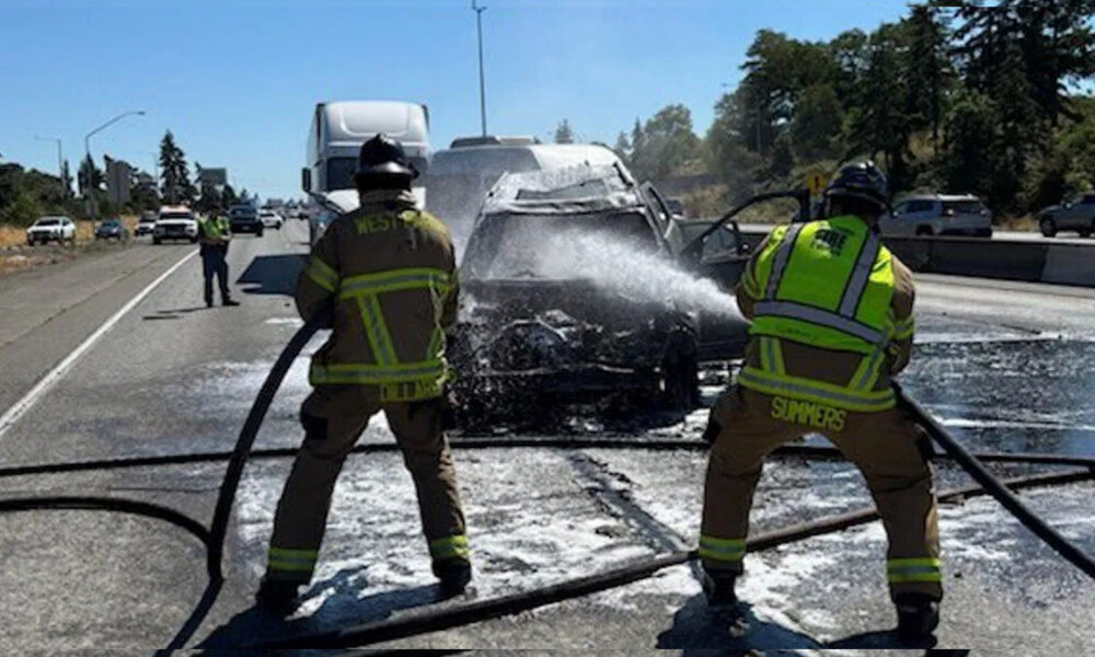 Firefighters in turnout gear spray water on a burning car on a highway, with a charred vehicle and traffic in the background.