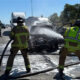 Firefighters in turnout gear spray water on a burning car on a highway, with a charred vehicle and traffic in the background.