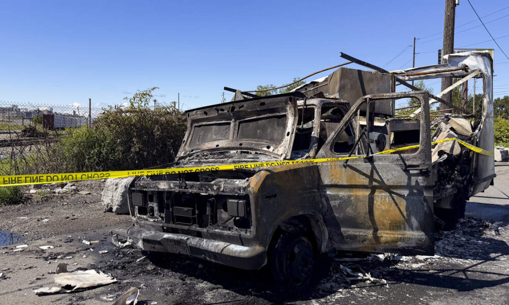 Burned-out van with charred frame and interior, taped off with yellow caution tape in an outdoor lot