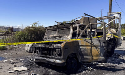 Burned-out van with charred frame and interior, taped off with yellow caution tape in an outdoor lot
