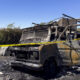 Burned-out van with charred frame and interior, taped off with yellow caution tape in an outdoor lot