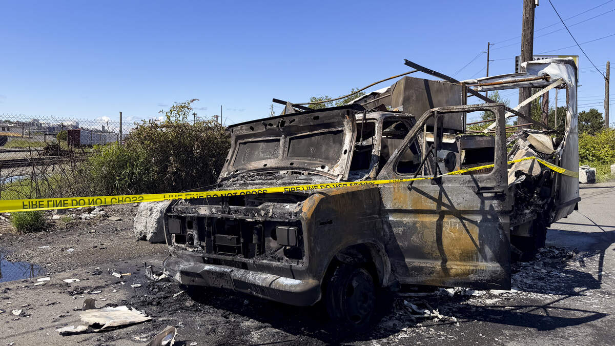 Burned-out van with charred frame and interior, taped off with yellow caution tape in an outdoor lot