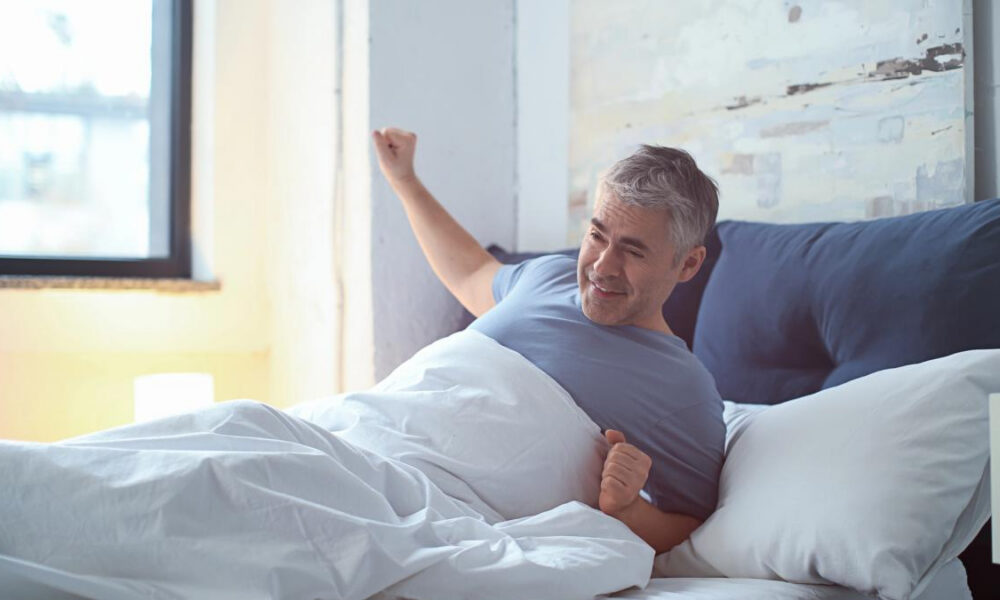 A smiling man in a blue shirt wakes up in bed and stretches under white sheets in a sunlit room.