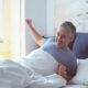 A smiling man in a blue shirt wakes up in bed and stretches under white sheets in a sunlit room.