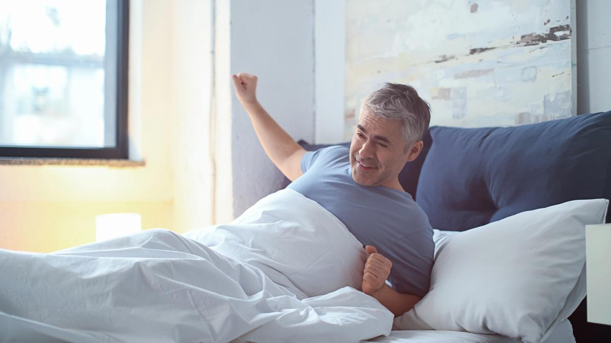 A smiling man in a blue shirt wakes up in bed and stretches under white sheets in a sunlit room.