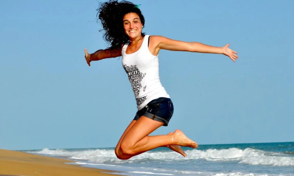 Woman in a white tank top and dark shorts jumps mid-air on a sandy beach, blue ocean behind her.