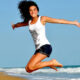Woman in a white tank top and dark shorts jumps mid-air on a sandy beach, blue ocean behind her.