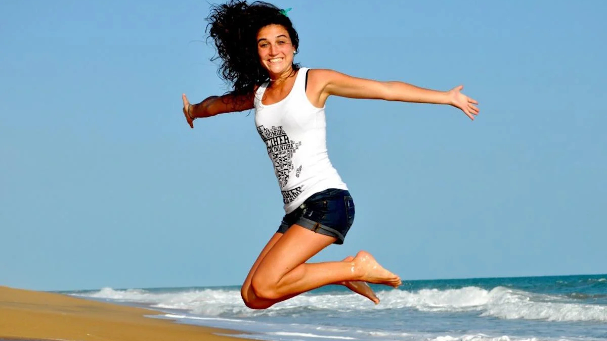 Woman in a white tank top and dark shorts jumps mid-air on a sandy beach, blue ocean behind her.