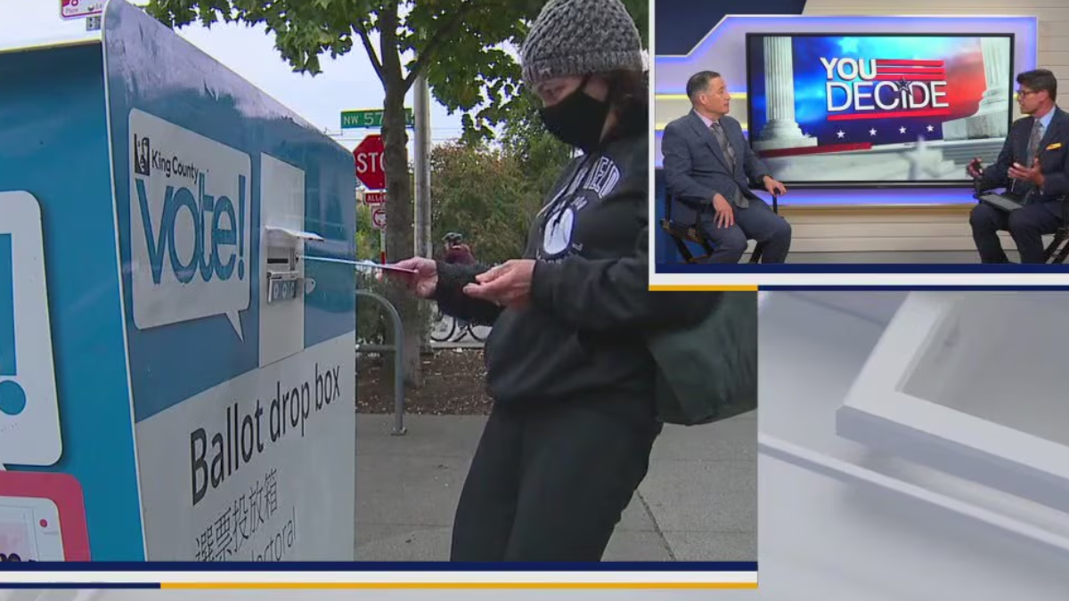 Voter in a knit cap and mask drops a ballot into a blue ballot drop box; inset shows a news set with 'YOU DECIDE'.