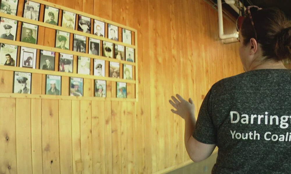 Woman in a Darrington Youth Coalition shirt stands before a wall of framed portrait photos mounted on wood panels.