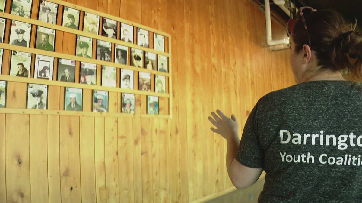 Woman in a Darrington Youth Coalition shirt stands before a wall of framed portrait photos mounted on wood panels.