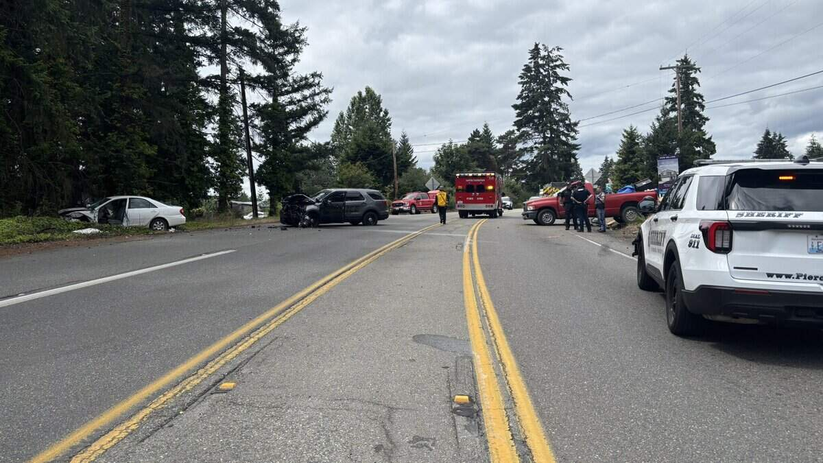 Two-lane road crash scene with a white car off the left shoulder, a darkened vehicle in the middle, and multiple emergency vehicles in the distance amid trees.